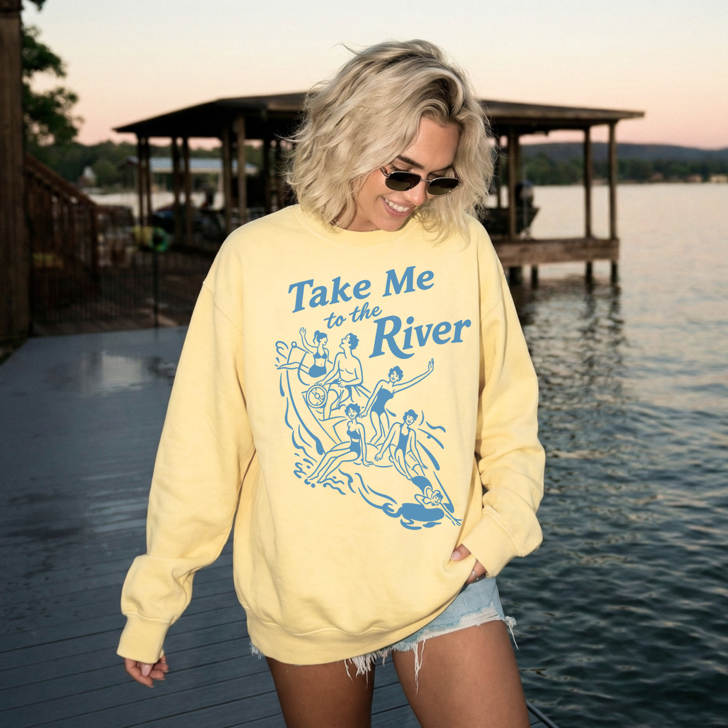 Yellow lake life sweatshirt with Take Me to the River graphic worn by woman near dock